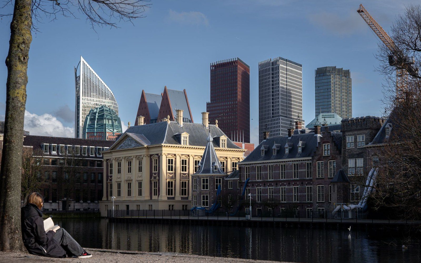 Person reading by a canal in The Hague, with historic buildings and skyscrapers behind