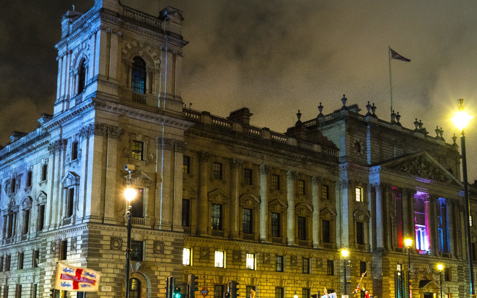A grand stone government building lit up at night with a Union Jack flying above