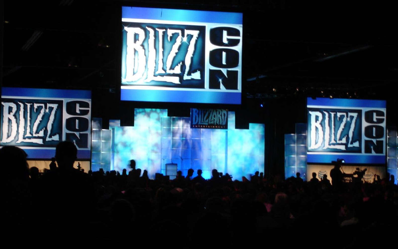 A developer on a large keynote stage mid-presentation, slide visible behind them showing a product roadmap, audience silhouettes in foreground